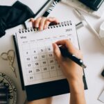 Top view of hands using a planner with coffee and accessories, focused on May 2021 calendar.
